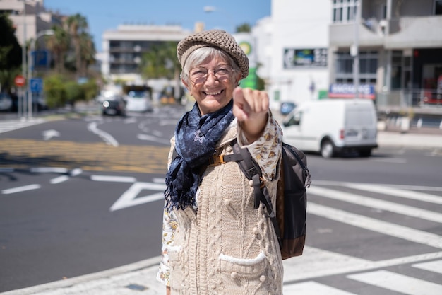 Feliz anciana viajera en el soleado centro de la ciudad expresando positividad buen humor sosteniendo mochila Atractiva anciana mirando a la cámara señalando con el dedo