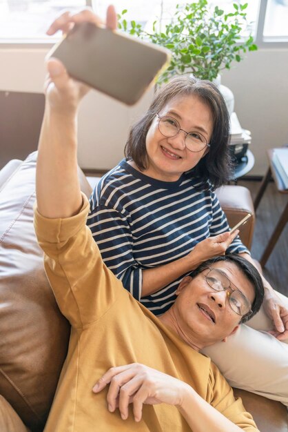 Feliz anciano amante asiático pareja sosteniendo teléfono inteligente mirando la pantalla del teléfono celular riendo casual relajante sentarse en el sofá juntos sonriendo ancianos abuelos maduros familia abrazando el estilo de vida