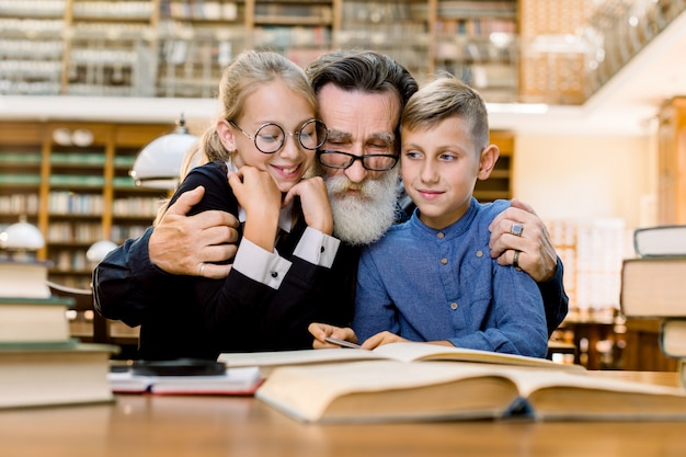 Feliz anciano barbudo, abuelo y su lindo nieto y nieta están sentados a la mesa en la antigua biblioteca vintage y leyendo libros