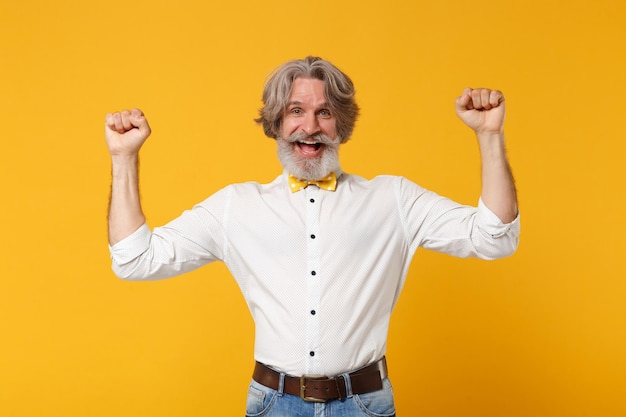 Feliz anciano de cabello gris bigote barbudo hombre con camisa blanca y corbata de moño posando aislado sobre fondo de pared naranja amarillo. Concepto de estilo de vida de las personas. Simulacros de espacio de copia. Apretando los puños como ganador.