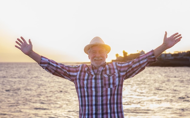 Feliz anciano caucásico con sombrero parado en la playa en la hora dorada de la puesta de sol mirando a la cámara con los brazos extendidos hombre barbudo disfrutando del tiempo libre de vacaciones o jubilación