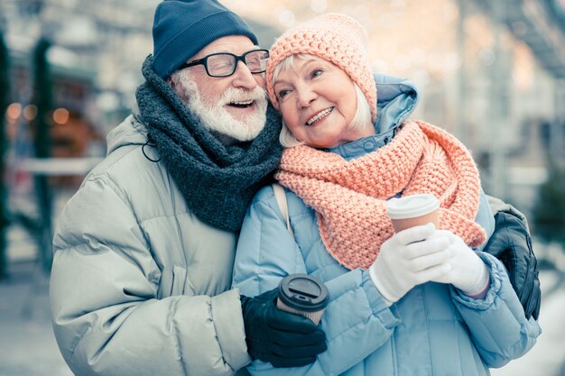 Feliz anciano cerrando los ojos y sonriendo mientras está al aire libre en un día de invierno con su esposa y abrazándola. Tazas de cartón de café en manos de los jubilados