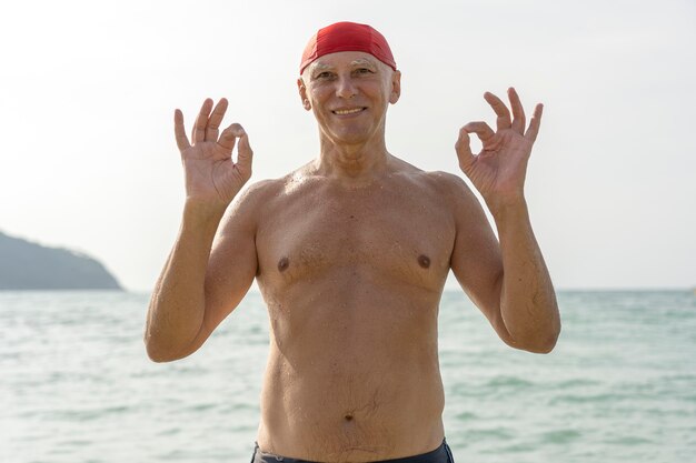 Feliz anciano con un gorro de baño rojo en la playa cerca del agua de mar