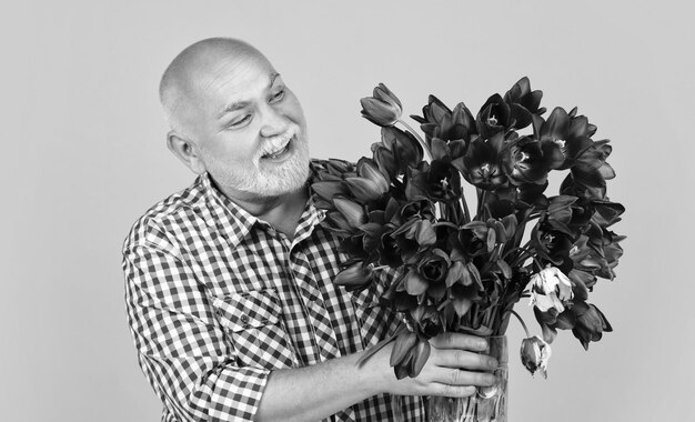 Feliz anciano jubilado con barba sostiene flores de tulipán de primavera sobre fondo amarillo