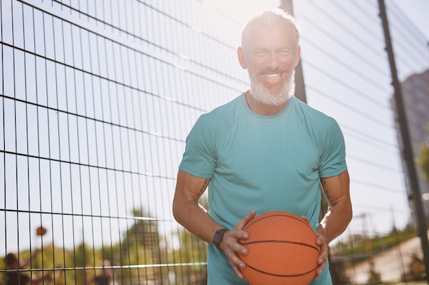 Feliz anciano jugador de baloncesto en ropa deportiva sosteniendo una pelota de baloncesto y sonriendo a la cámara mientras