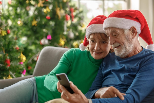 Feliz anciano y mujer madura con sombreros de Santa sonriendo y navegando por las redes sociales en el teléfono celular juntos mientras están sentados en el sofá cerca del árbol de Navidad