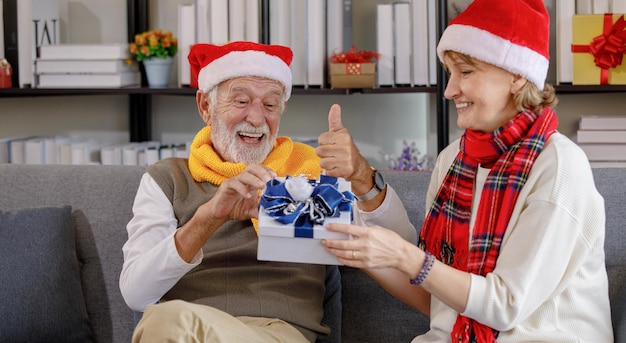 Feliz anciano riendo mientras está sentado en el sofá y recibe el presente de la esposa madura en bufanda y gorro de Papá Noel durante la celebración de Navidad en casa