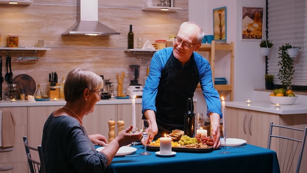 Feliz anciano sirviendo a su esposa con uvas y queso. Pareja de ancianos hablando, sentados a la mesa en la cocina, disfrutando de la comida, celebrando su aniversario en el comedor