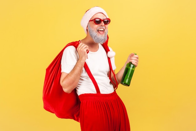 Feliz anciano con sombrero de santa claus, gafas de sol de moda y pantalones con tirantes sosteniendo una botella con alcohol y una gran bolsa roja, celebración, mal hábito. Disparo de estudio interior aislado sobre fondo amarillo