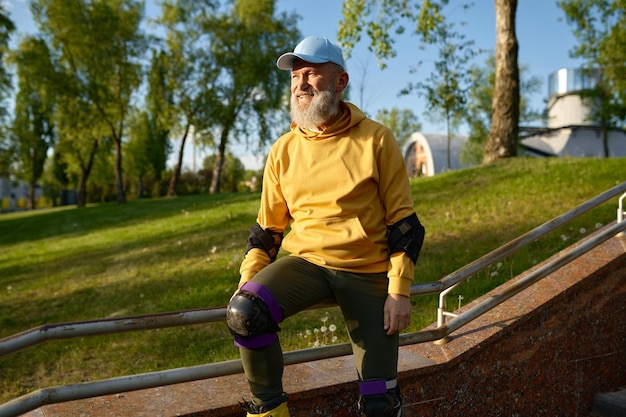 Feliz anciano sonriente con patines descansa sentado en la baranda de la escalera en el parque de la ciudad. Tiempo activo en la jubilación