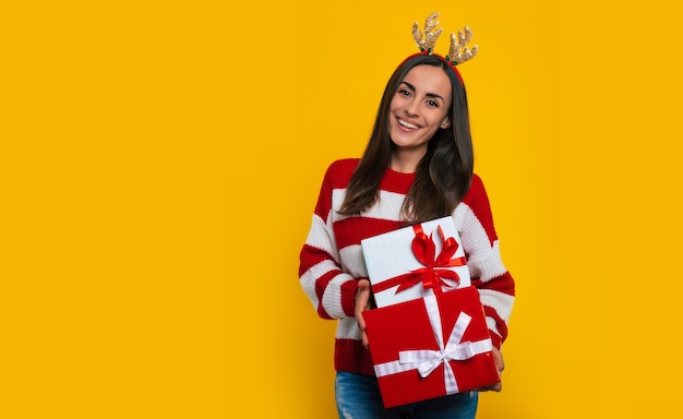 Feliz animada jovem morena com chapéu de veado, com muitas caixas de presente de Natal coloridas nas mãos isoladas em fundo amarelo.