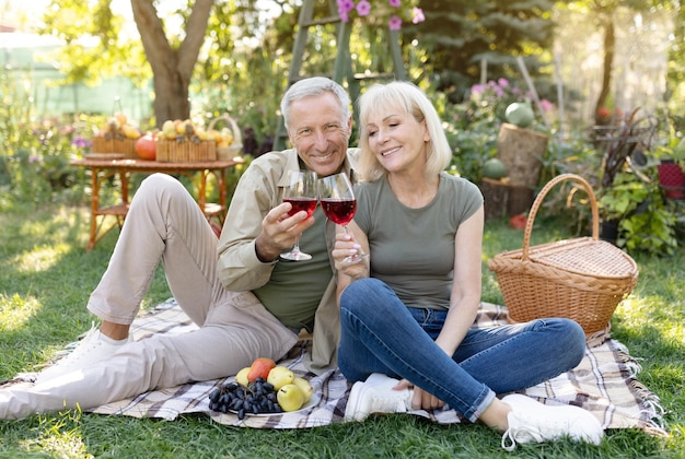 Feliz aniversario amorosa pareja mayor casada bebiendo vino mientras se sienta en una manta haciendo un picnic