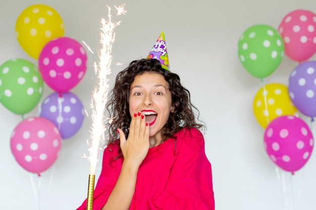 Feliz aniversário. Garota morena sexy posando com balões, fogos de artifício, balões coloridos e bolo de férias em um fundo branco