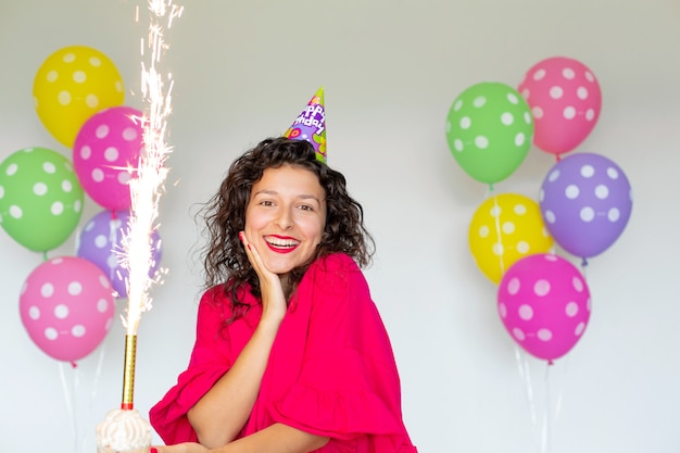 Feliz aniversário. Garota morena sexy posando com balões, fogos de artifício, balões coloridos e bolo de férias em um fundo branco