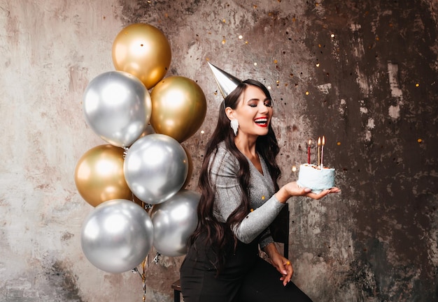 Feliz aniversário morena sexy posando com balões bolo de aniversário de fogos de artifício no fundo de uma parede decorativa uma mulher sopra as velas no bolo