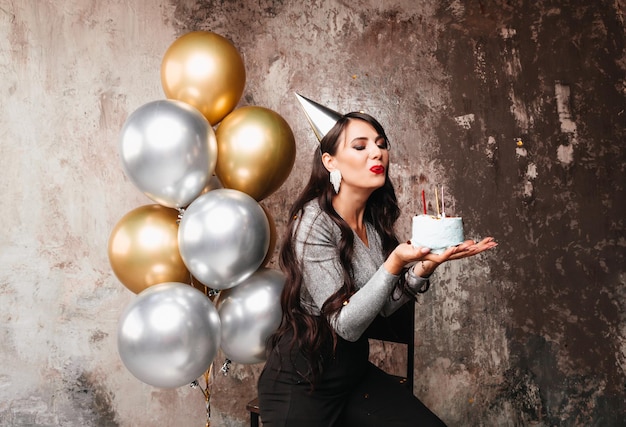 Feliz aniversário morena sexy posando com balões bolo de aniversário de fogos de artifício no fundo de uma parede decorativa uma mulher sopra as velas no bolo