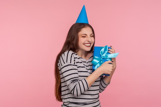 Feliz aniversário para mim. Alegre mulher encantada com chapéu de cone de festa abraçando a caixa de presente embrulhada e sorrindo com emoção, regozijando-se presente, celebração do feriado. tiro de estúdio isolado no fundo rosa