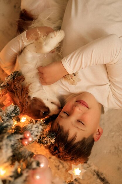 Foto feliz ano novo um menino em pijama caseiro leve abraça seu animal de estimação cavalier king charles spaniel em casa no quarto perto da árvore de natal