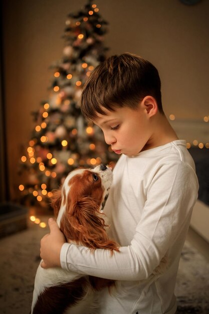 Foto feliz ano novo um menino em pijama caseiro leve abraça seu animal de estimação cavalier king charles spaniel em casa no quarto perto da árvore de natal