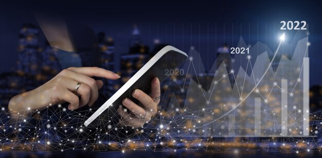 Feliz año nuevo 2022 - tableta blanca táctil de mano con signo de gráfico de gráfico de crecimiento de holograma digital sobre fondo borroso oscuro de la ciudad. Gráficos financieros que muestran ingresos crecientes en 2022