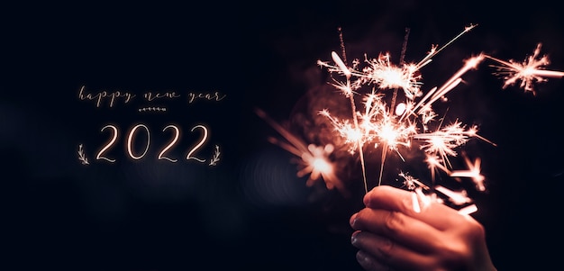 Feliz año nuevo 2022 texto con mano sosteniendo ardiente explosión de fuegos artificiales de bengala con un fondo negro bokeh en la noche, fiesta de evento de celebración navideña, tono vintage oscuro