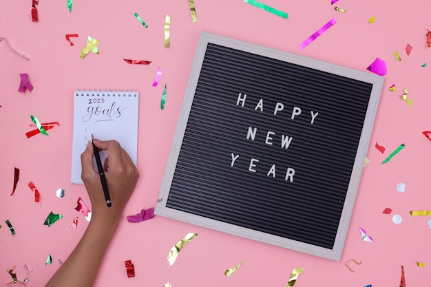 Feliz Año Nuevo 2023. Mano de mujer escribiendo 2023 Metas en un cuaderno decorado con partículas de confeti o