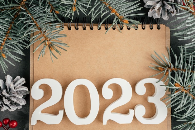 Feliz Año Nuevo 2023 Números blancos 2023 acostados en un cuaderno doblado con ramas de árboles de Navidad y piñas copian espacio