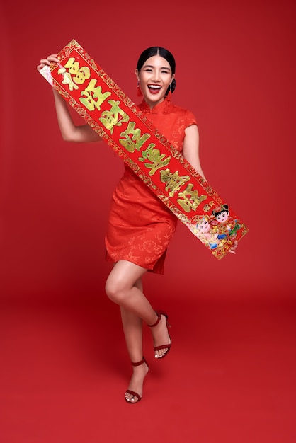 Feliz Año Nuevo Chino. Mujer asiática con vestido tradicional cheongsam qipao sosteniendo coplas de año nuevo chino aisladas sobre fondo rojo.