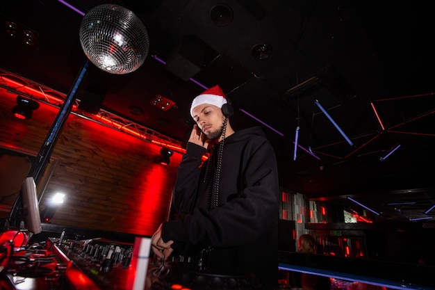 Feliz año nuevo y feliz Navidad carismático disc jockey con sombrero rojo de santa claus, auriculares y sudadera con capucha reproduce música en tocadiscos de DJ. Concepto de fiesta de año nuevo.
