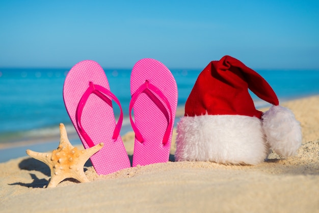 Feliz año nuevo en el mar. Sandalias y gorro de Papá Noel en la playa de arena - concepto de vacaciones de Navidad