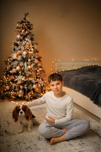 Feliz año nuevo un niño en pijama casero ligero abraza a su mascota Cavalier King Charles Spaniel en casa en el dormitorio cerca del árbol de Navidad