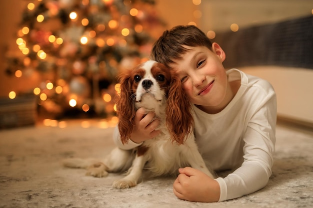 Feliz año nuevo un niño con pijamas caseros ligeros abraza a su mascota cavalier king charles spaniel en casa en el dormitorio cerca del árbol de Navidad
