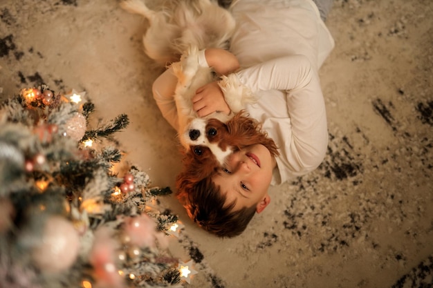 Feliz año nuevo un niño con pijamas caseros ligeros abraza a su mascota cavalier king charles spaniel en casa en el dormitorio cerca del árbol de Navidad