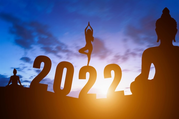 Feliz año nuevo números 2022, silueta estatua de Buda y silueta mujer practicando yoga amanecer temprano en la mañana sobre el fondo del horizonte, concepto de feliz año nuevo.