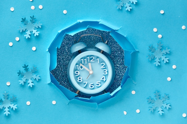 ¡Feliz año nuevo! Reloj de brazo en agujero de papel, vista superior plana sobre papel azul con nieve y copos de nieve