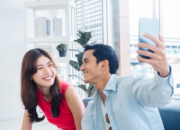 Feliz apuesto hombre asiático y joven hermosa mujer selfie mientras se sienta cerca de la ventana de cristal en la sala de estar blanca en un edificio alto Pareja alegre tomando selfie de retrato con teléfono móvil inteligente