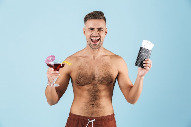 Feliz apuesto joven sin camisa con pantalones cortos de playa de pie sobre azul, bebiendo cócteles, mostrando el pasaporte con boletos