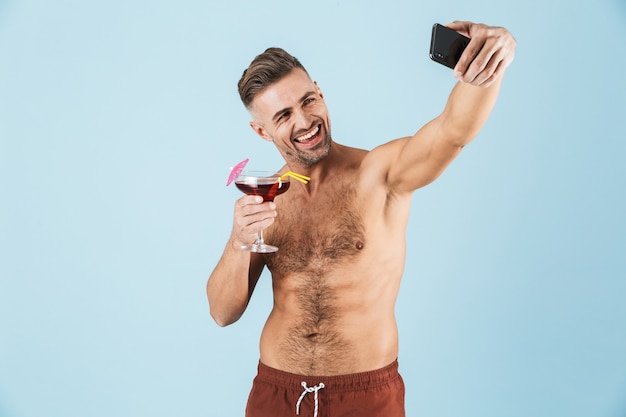 Feliz apuesto joven sin camisa con pantalones cortos de playa de pie sobre azul, tomando un selfie mientras bebe un cóctel