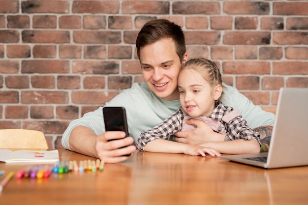 Feliz apuesto joven padre con tatuaje sentado a la mesa en loft y tomando selfie con hija para enviar una foto a la madre