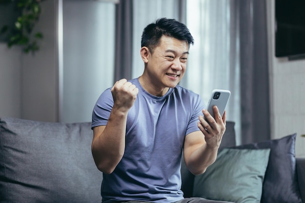 Feliz asiático en casa sentado en el sofá disfrutando de escribir un mensaje de teléfono hombre alegre y sonriente en la sala de estar del loft