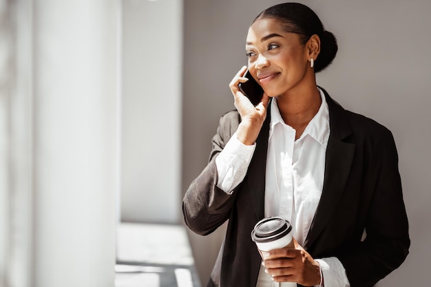 Feliz y atractiva empresaria negra tomando café para llevar y hablando por teléfono en la oficina