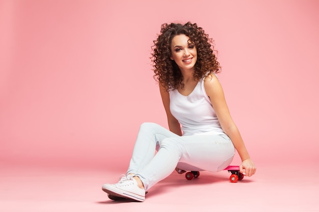 Feliz atractiva joven sentada en patineta sobre rosa.