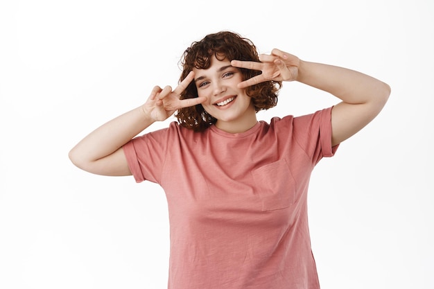 Feliz y atractiva mujer joven que muestra un gesto kawaii de paz con el signo v cerca de los ojos, baila discoteca, sonríe y expresa positividad con alegría, de pie sobre fondo blanco