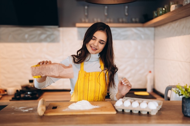 Feliz atractivo joven adulto mujer señora ama de casa panadero usar delantal espolvorear harina sobre la mesa de la cocina para hornear pasteles.