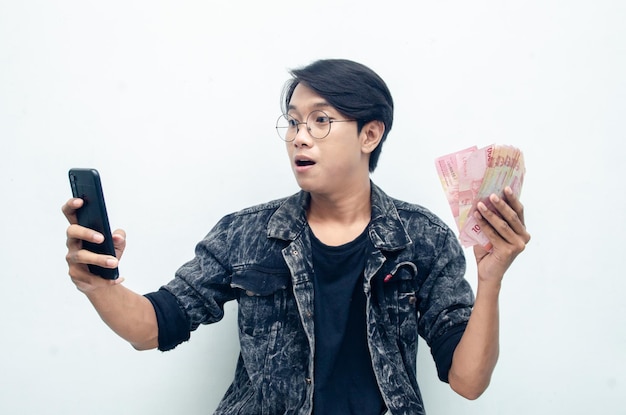 Feliz y atractivo joven indonesio sorprendido felizmente mientras sostenía papel moneda rupia y teléfono.