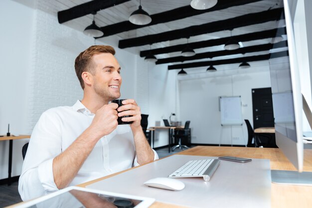 Feliz atraente jovem empresário sentado e bebendo café no local de trabalho