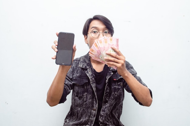 Feliz atraente jovem indonésio chocado alegremente enquanto segurava o papel-moeda e o telefone da rupia.