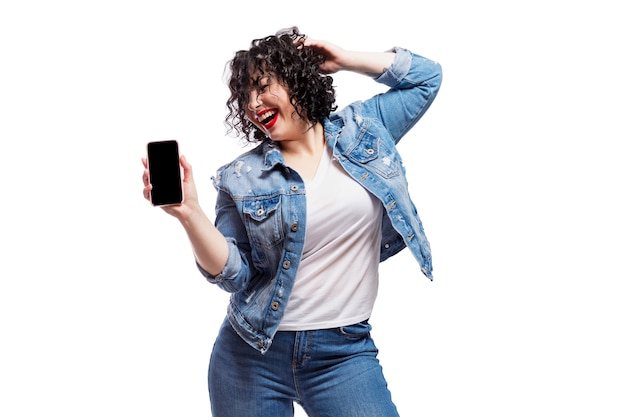 Feliz bailando joven bella mujer en jeans muestra un teléfono inteligente que se emite en una pantalla en negro. Morena brillante con el pelo rizado.
