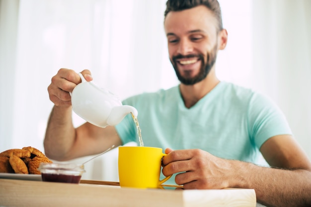 Feliz barbudo jovem está sentado em sua cama tomando café com chá ou café