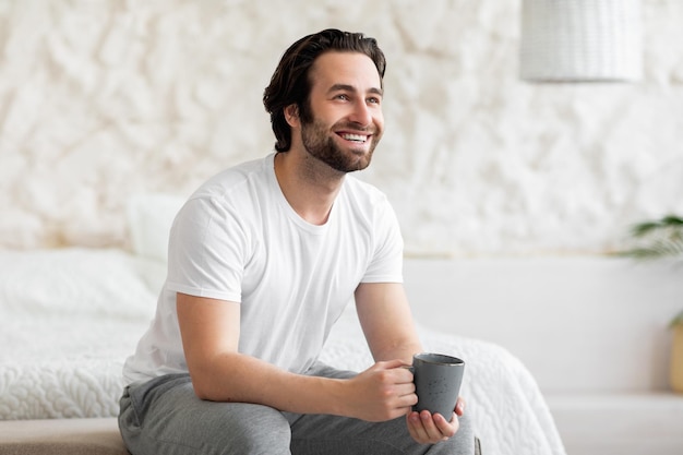 Feliz barbudo jovem sentado na cama tomando café